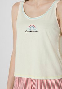 Débardeur jaune clair avec des bordures bleues et rose, présentant un motif dearc-en-ciel brodé avec le texte "Au-delà de l'arc-en-ciel" en dessous.