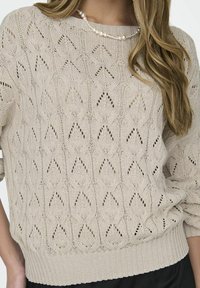 Sweter o splocie w jasnym beżu z teksturowanym wzorem w diamenty, ściągaczami przy mankietach i dolnym brzegu. Zaprojektowany w luźnym kroju i z dekoltem typu łódka.