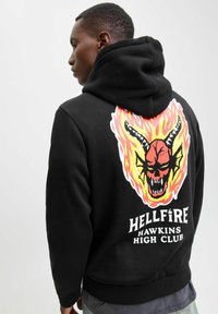 Schwarzer Hoodie mit einem lebhaften Grafikdesign eines Schädels mit Flammen, Hörnern und dem Schriftzug "HELLFIRE HAWKINS HIGH CLUB" auf der Rückseite.