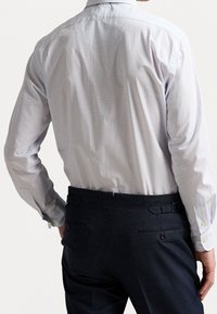 Man som bär en ljus mönstrad långärmad skjorta instoppad i mörka skräddarsydda byxor med justerbara sidoflikar, sedd bakifrån.