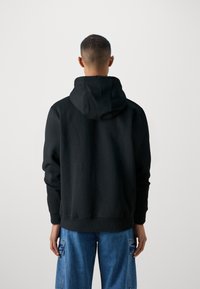 Uomo che indossa un felpa con cappuccio nera e jeans cargo blu, girato di spalle su uno sfondo chiaro e semplice.