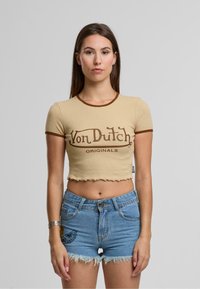 Bež kratka majica s rjavim trimom, ki prikazuje logo "Von Dutch Originals". Kombinirana s svetlo modrimi kavbojkami s strganim robom.