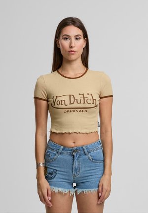 Beige Cropped-T-Shirt mit braunem Saum, mit dem "Von Dutch Originals"-Logo. Kombiniert mit hellblauen Denim-Shorts mit fransigem Saum.