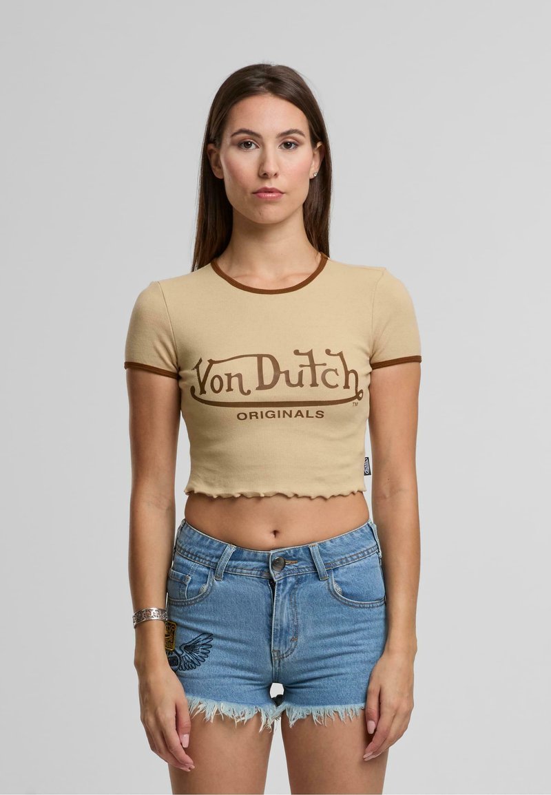 Bež kratka majica s rjavim trimom, ki prikazuje logo "Von Dutch Originals". Kombinirana s svetlo modrimi kavbojkami s strganim robom.
