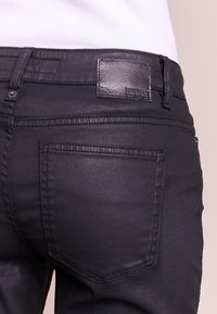Jean en denim noir avec une texture lisse, featuring un patch en cuir à la taille et des détails de couture subtils sur les poches arrière.