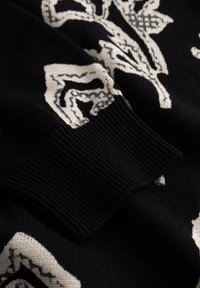 Crni pleteni džemper s bijelim cvjetnim uzorkom. Ima rebrasti rub i glatku teksturu, naglašavajući kontrast između boja.