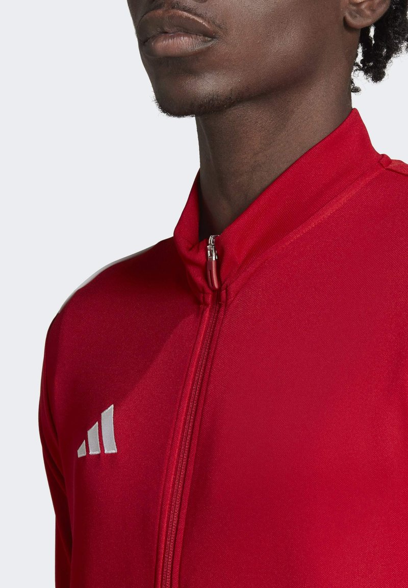 Chaqueta roja con cierre y alto collar, hecha de un tejido suave. Presenta un logo plateado en el lado izquierdo del pecho y rayas laterales en contraste.