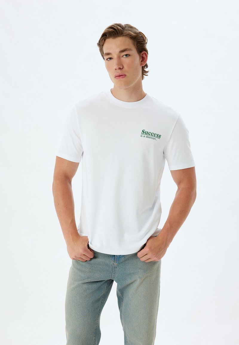 Hvid bomulds-t-shirt med grøn tekst "Succes er en beslutning." Klassisk crewneck-design, korte ærmer og afslappet pasform.