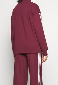 Chándal burdeos con mangas largas, cuello alto y rayas blancas en los pantalones. Tejido suave, corte relajado y detalles de costura visibles.