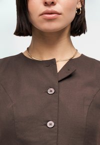 Γυναίκα που φοράει καφέ μπλούζα με κουμπιά, χρυσό κολιέ και χρυσούς κρίκους στα αυτιά, με κοντά σκούρα μαλλιά, εμφανίζοντας τον λαιμό και το κάτω μέρος του προσώπου.