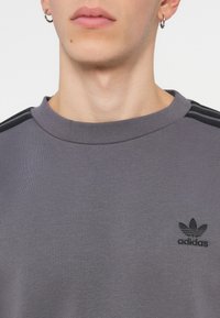 Grå sweatshirt i strukturerad tyg med rund nacke och svarta tre-strimmiga detaljer på axlarna. Inkluderar liten svart logotyp.