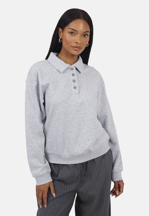 Vrouw met lang zwart haar, zilveren oorringen, een lichtgrijze trui met knopen aan de kraag en donkergrijze broek met fijne krijtstrepen.