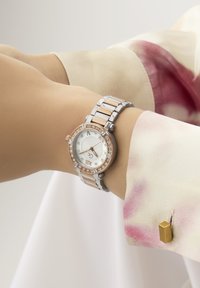 Reloj plateado y de oro rosa con una esfera circular, acentos de diamantes y una pulsera de metal bicolor. Lleva puesto en una mano con una blusa en tonos pastel.