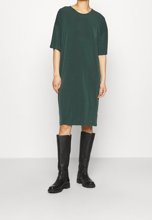 Personne vêtue d'une robe ample vert foncé longueur genou avec manches courtes et bottes en cuir noires montant jusqu'aux genoux, debout devant un fond uni.