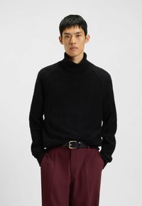 Schwarzer, strukturierter Rollkragenpullover mit geripptem Kragen. Getragen mit burgunderroten Hosen und einem schwarzen Gürtel mit silberner Schnalle.