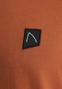 Tessuto di camicia marrone con una texture liscia presenta una toppa rettangolare nera con un motivo astratto bianco.