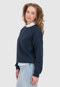 Felpa blu navy cropped con polsini a costine e scollatura a girocollo, indossata sopra una camicia bianca con colletto. Design semplice, texture morbida.
