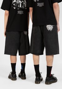 Des shorts en denim noir présentent des poches arrière distinctes avec des motifs brodés blancs, associés à des chaussettes noires et des chaussures noires.