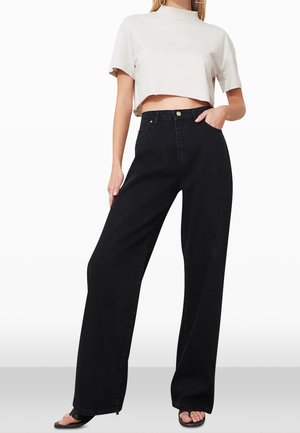 Kobieta ubrana w czarne jeansy o szerokich nogawkach, biały krótki top z krótkim rękawem i czarne sandały z odkrytymi palcami, stoi z jedną ręką w kieszeni.