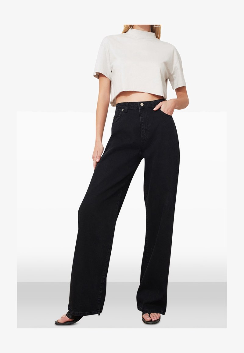 Femme portant un jean noir à jambes larges, un top court blanc à manches courtes et des sandales noires à bouts ouverts, debout avec une main dans la poche.