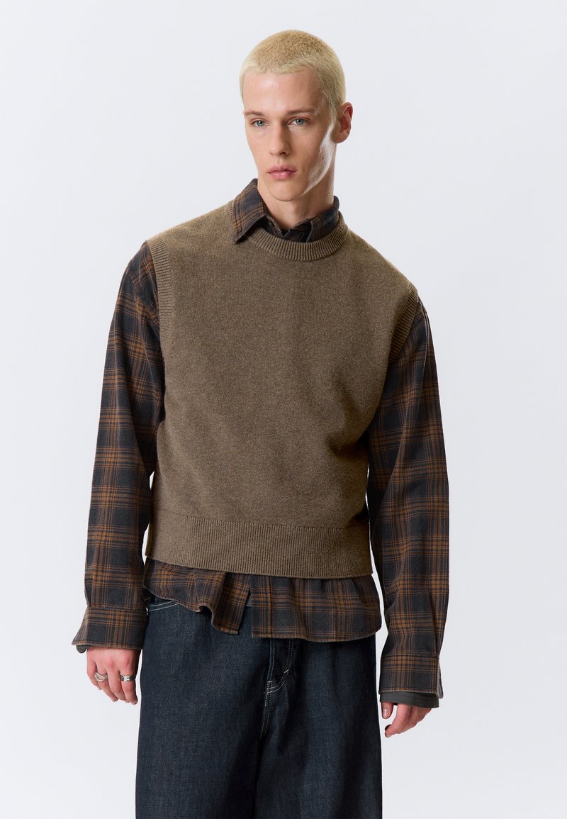 Hnedá pletená vesta cez tmavú károvanú košeľu s dlhým rukávom v odtieňoch hnedej a sivej. Hnedé denimové nohavice s mierne rozšírenými nohavicami.