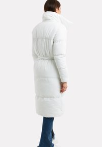Lang hvid polstret frakke med høj krave, bælte i taljen og lodret syning. Stoffet har en glat tekstur og en fyldig silhuet.