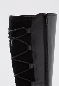 Bota negra hasta la rodilla que combina cuero suave y ante blando. Incluye un zipper lateral y detalle de cordones a lo largo del tubo.