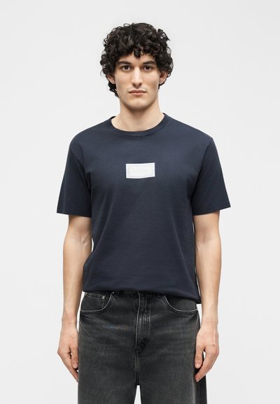 Άνδρας με σγουρά σκούρα μαλλιά στέκεται εμπρός, φοράει απλό σκούρο μπλουζάκι με μικρό ορθογώνιο λευκό ταμπελάκι στο στήθος και ξεβαμμένο μαύρο τζιν.