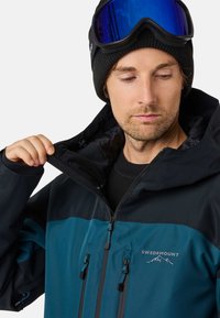 Skijas met een zwart en teal kleurblokontwerp, gemaakt van waterdicht materiaal, met verstelbare koordjes en een logo.