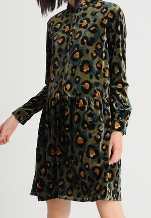 Mujer con un vestido de manga larga hasta la rodilla con estampado de leopardo en verde, negro y naranja y una cremallera frontal.