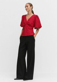 Kobieta w teksturowanej czerwonej bluzce z dekoltem w kształcie litery V i bufkami oraz w szerokich czarnych spodniach, stojąca z rękami w kieszeniach na białym tle.