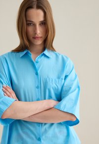 Camisa de botones de manga corta en azul claro con cuello puntiagudo, un bolsillo en el pecho y una tela ligeramente texturizada. Los botones combinan con el color de la camisa.