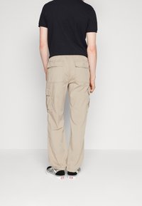 Beige cargo kalhoty s několika kapsami, elastickým pasem a volným střihem, v kombinaci s černobílými teniskami na jednobarevném pozadí.