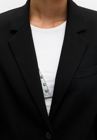 Blazer noir avec des revers nets et un seul bouton. En dessous, une chemise blanche présente un panneau en denim bleu en forme de V avec des accessoires argentés.