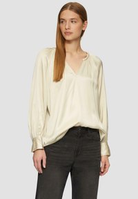 Blouse en crème légère et soyeuse avec un col en V, des épaules plissées et des manches longues se terminant par des poignets en satin, associée à un jean gris foncé.