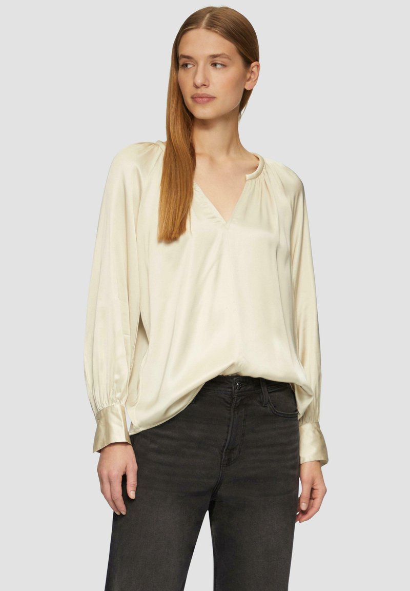 Blouse en crème légère et soyeuse avec un col en V, des épaules plissées et des manches longues se terminant par des poignets en satin, associée à un jean gris foncé.