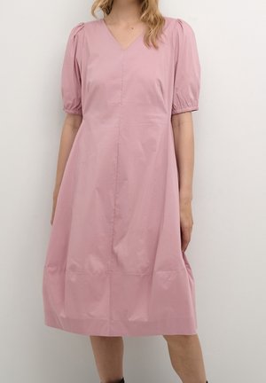 Frau, die ein knielanges, hellrosa Kleid mit puffärmeligen kurzen Ärmeln und einem V-Ausschnitt trägt, steht vor einer schlichten hellen Wand.