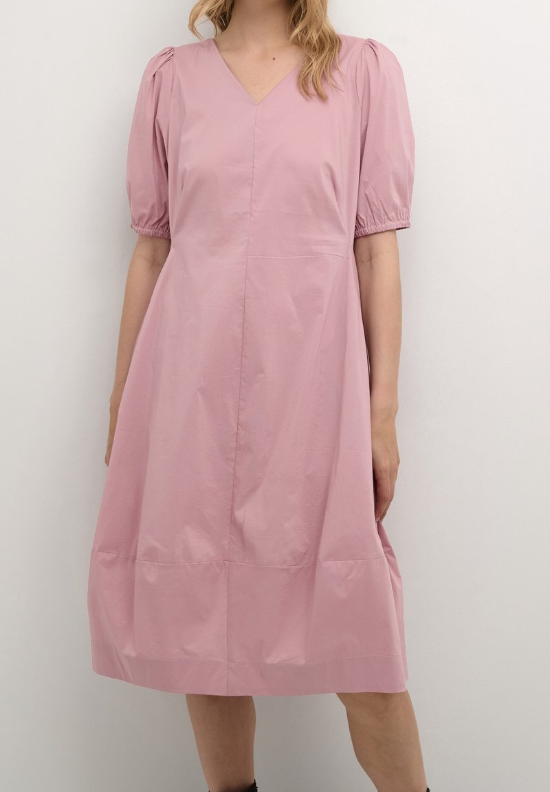 Kvinna klädd i en knälång ljusrosa klänning med puffärmar och V-hals som står mot en enkel ljus vägg.