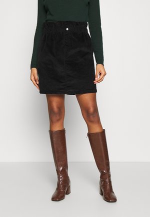 Falda mini de pana negra con cinturilla elástica, combinada con botas marrones brillantes hasta la rodilla, con acabado texturizado y tacones cuadrados.