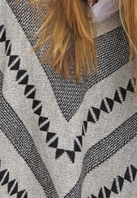 Długie jasnobrązowe włosy opadają na szary wełniany sweter z czarnymi geometrycznymi wzorami w trójkąty oraz widocznym białym kołnierzykiem.