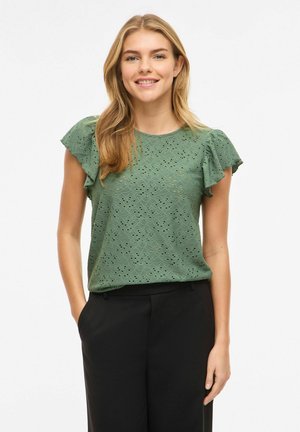 Fiatal nő hosszú, szőke hajjal, mosolyogva, zöld tavaszi csipkés rövid ujjú blúzt és fekete nadrágot viselve, egyik kezét a zsebében tartva áll.