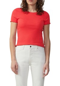 Vrouw in een nauwsluitend kort mouw ribgebreid rood top en hoog-waist witte broek, staand tegen een witte achtergrond.