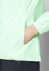 Light green lightweight jacket features a textured pattern, elastic cuffs, and side pockets. Logo tag visible at the lower seam.