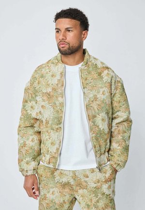 Un homme portant une veste et un pantalon floral à motifs verts et beiges, avec une chemise blanche, se tenant avec une main dans sa poche.