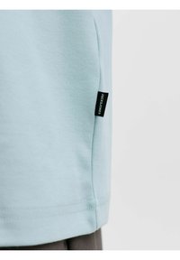 Hellblaues Baumwoll-T-Shirt mit kurzen Ärmeln, das einen abgesteppten Saum und ein schwarzes Etikett auf der linken Seite aufweist. Glatte Textur.