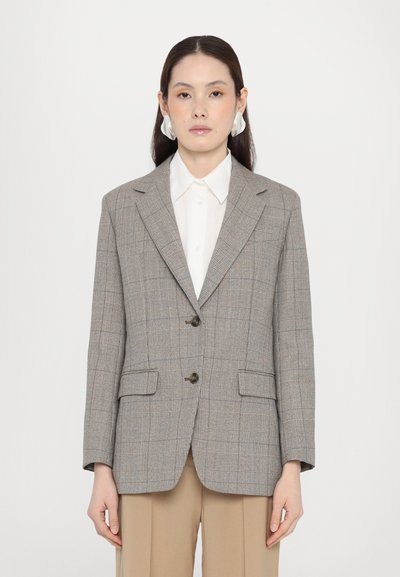 Blazer grigio a quadri con due bottoni, rever a scallop e tasche frontali. Indossato su una camicia bianca, abbinato a pantaloni beige.