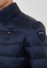 Primo piano di una persona che indossa un giubbotto imbottito Blauer U.S.A. di colore blu navy con colletto alto e una toppa con logo sulla manica.