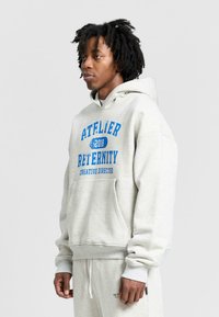 Jasnoszara bluza z kapturem z niebieskim nadrukiem "ATELIER 2010 RETERNITY CREATIVE DIRECTOR." Posiada kieszeń kangurkową i ściągacze przy rękawach.