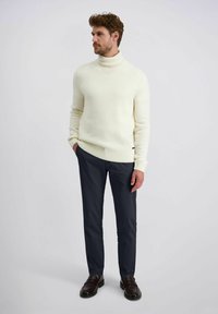 Man in een witte coltrui, marineblauwe slim-fit broek en donkerbruine instappers, staande met zijn handen in zijn zakken en kijkend naar rechts.