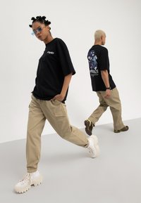 Zwei Modelle tragen schwarze T-Shirts mit Grafiken und beige Cargo-Hosen und posieren dynamisch vor einem schlichten hellen Hintergrund.